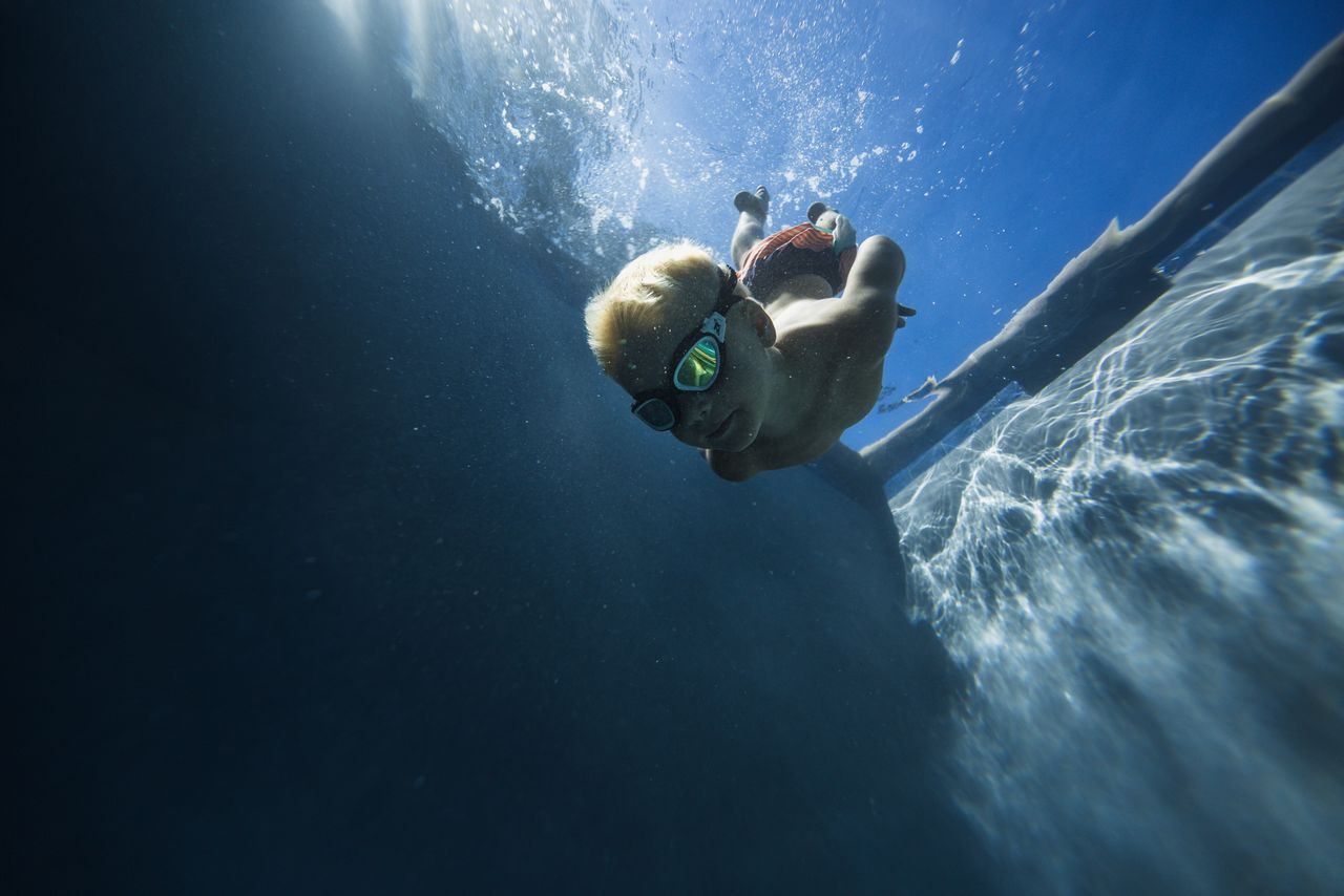 A young boy wearing goggles dives underwater in a sunny pool, embodying summer fun.